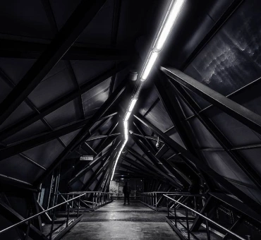 Futuristic holl-way in dark black-and-gray colors with metal balustrade and long lamp through the whole ceiling and standing man in the end of it.